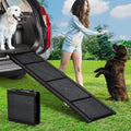 Four-panel folding aluminium dog ramp with carpeted tread running from grass to an SUV boot, golden retriever at the top and a chocolate labrador waiting